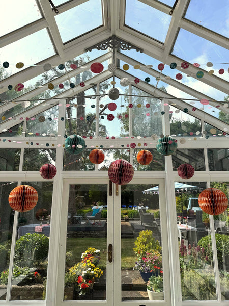 Colourful hanging honeycomb paper lanterns and circular garlands decorate a glass conservatory with white-framed windows and doors, overlooking a garden patio area.