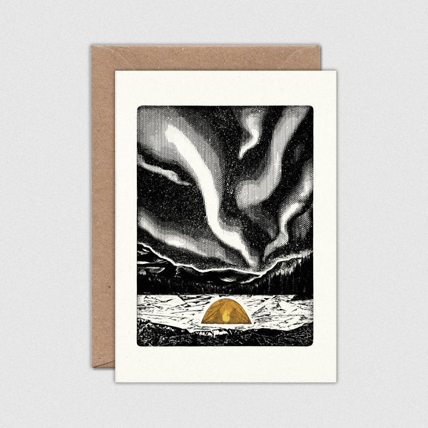 A greeting card with a kraft envelope behind it, featuring a black-and-white northern lights landscape with a small orange dome tent at the bottom.