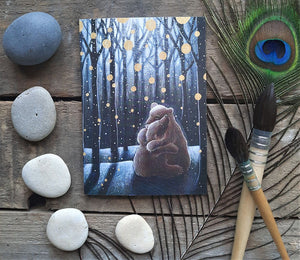 Card with bear illustration in a forest scene on a wooden surface with pebbles and a peacock feather.