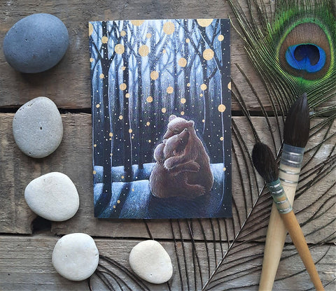 Card with bear illustration in a forest scene on a wooden surface with pebbles and a peacock feather.