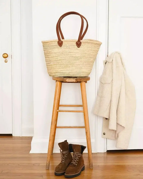 Woven basket on a wooden stool with brown boots and a beige coat against a white wall.