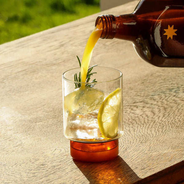 Brown bottle pouring liquid into a glass with ice and lemon slices on a wooden surface.