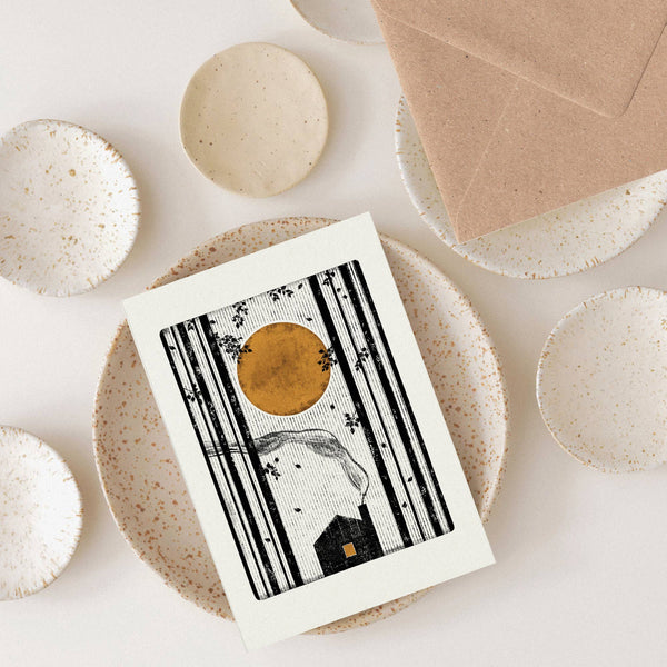 Artistic greeting card with a stylized house and sun on a textured surface with ceramic plates and a brown envelope.