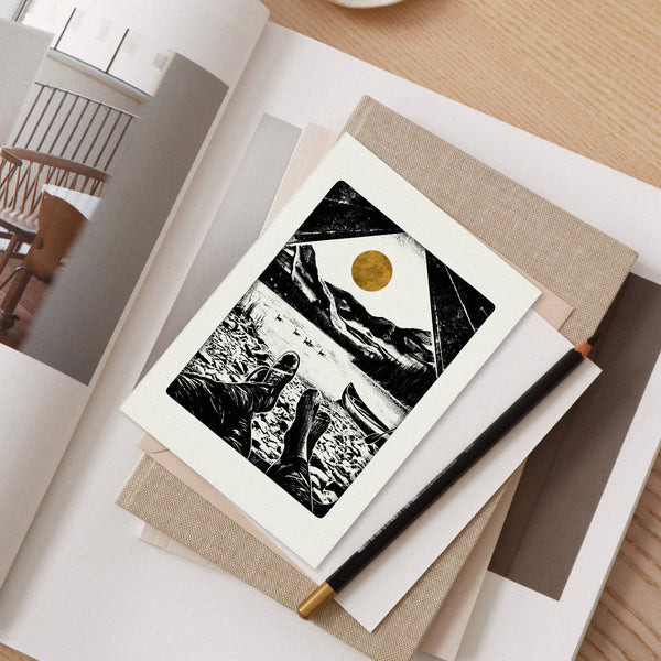 Artistic greeting card with a black and white mountain scene and gold sun, placed on a notebook with a pen.