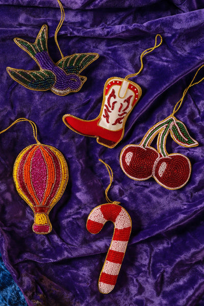 Collection of beaded decorations including a cowboy boot, hot air balloon, candy cane, and cherries on a purple fabric background.