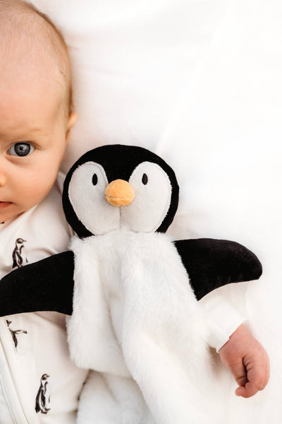 A close-up lifestyle shot of a baby lying on a white pillow, holding a plush penguin toy with black and white fur and a small orange beak.