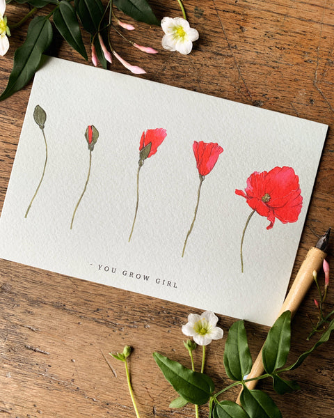 Card with red floral illustrations and 'You Grow Girl' text on a wooden surface with flowers and a pen.