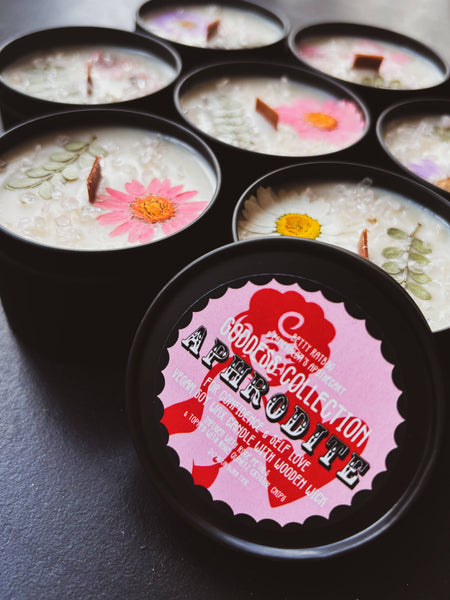 Candle tins with floral designs on a dark surface, featuring a label for 'Aphrodite'.