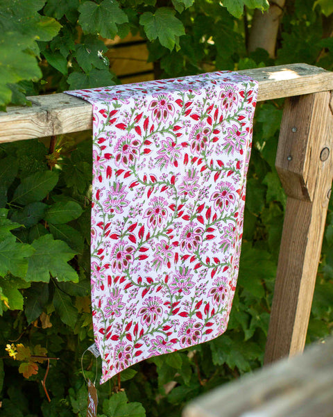 A tea towel featuring a pink and red floral pattern on a white background, draped over a rustic wooden fence post with green garden foliage in the background.