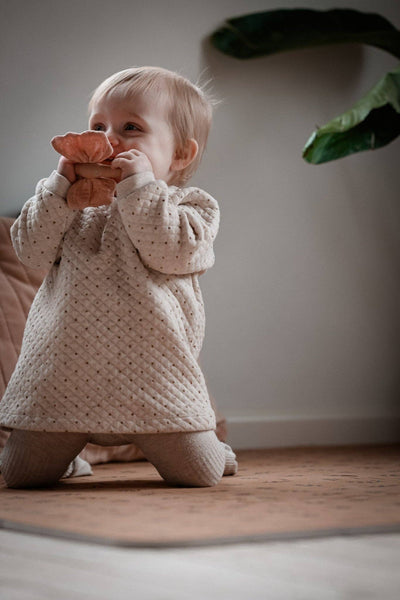 Baby in a beige knitted dress playing with a toy on a wooden floor.