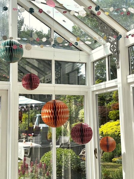 Colourful hanging honeycomb paper lanterns in orange, burgundy, and teal suspended inside a bright white conservatory with large windows and a garden view.