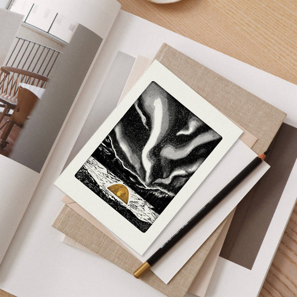 A greeting card with a white border featuring a black and white abstract aurora-like illustration and a small orange tent near the bottom, styled on top of stacked neutral books and papers with a black pencil.