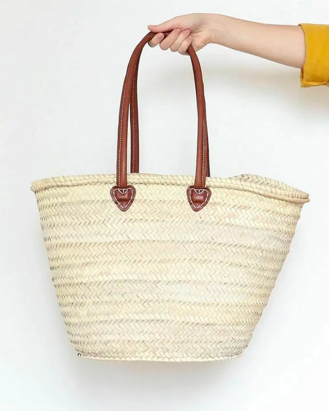 Woven straw tote bag with brown handles held by a hand on a white background