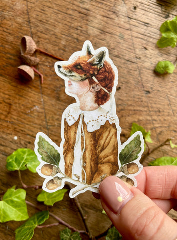 Sticker of a girl wearing a fox mask held by a hand on a wooden surface with leaves.