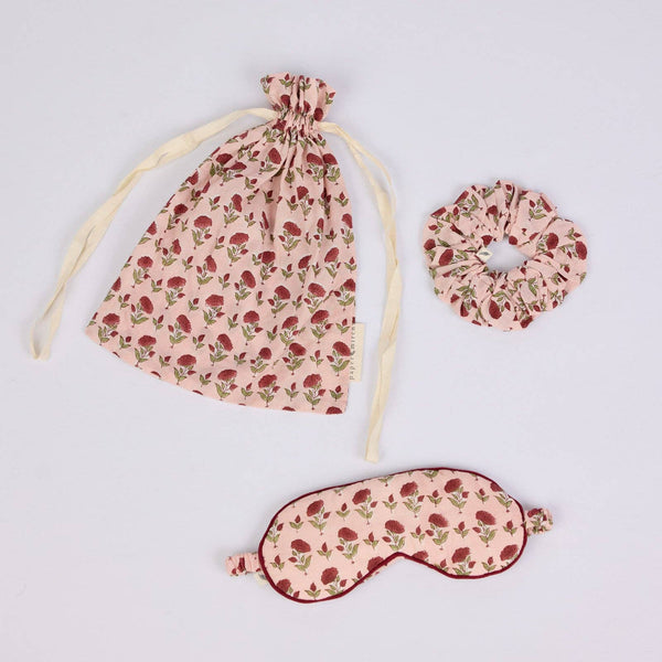 Three-piece sleep set featuring a drawstring bag, a scrunchie, and an eye mask in a dusty pink fabric with a red rose print, arranged on a light grey background.