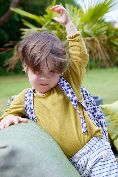 Child in a yellow shirt wearing a backpack sitting outdoors with greenery in the background
