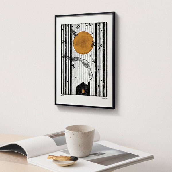 Framed artwork with a stylized illustration of a house and sun on a white wall above a table with a cup and magazine.