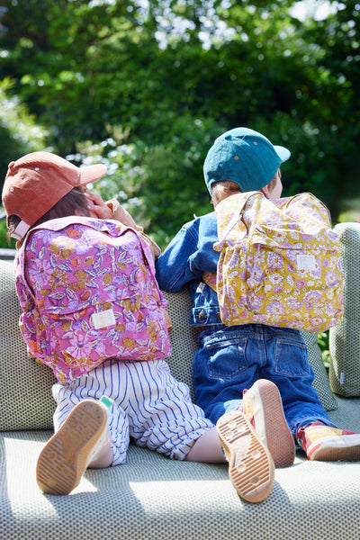 Two children sitting on a bench with colorful backpacks, surrounded by greenery.