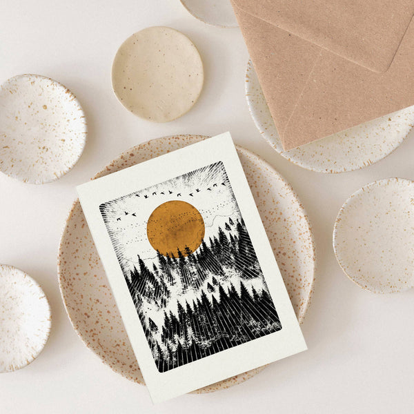 Greeting card with a sunset over trees design on a white surface with ceramic bowls.