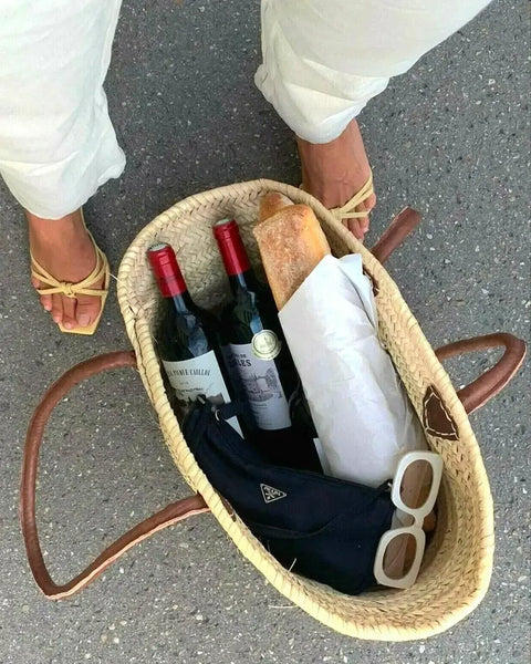 Wicker picnic basket with wine bottles, bread, and sunglasses on a concrete surface.