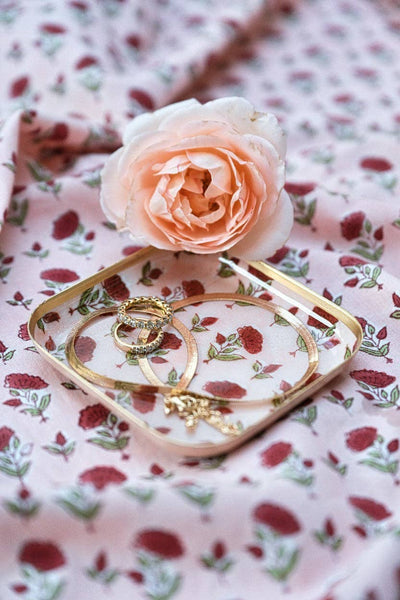 Gold rings and bracelets displayed on a clear square tray with a gold rim, styled with a peach rose on a pink floral fabric background.