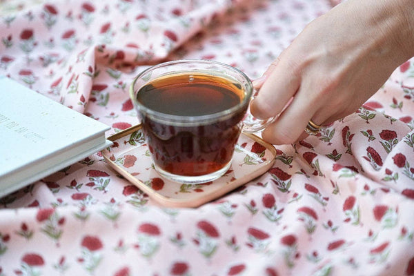 A hand holding a clear glass cup filled with dark tea, resting on a square coaster over a pink floral blanket, with a book partially visible on the left.