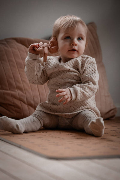 Baby in a textured sweater and pants sitting on a wooden floor with pink cushions in the background.