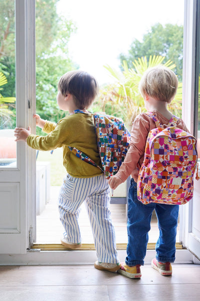 Two children with colourful backpacks standing by a door, ready to go outside.