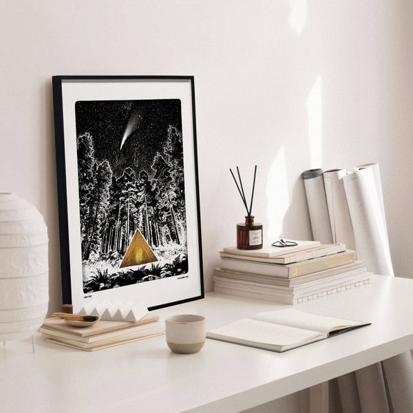 Framed artwork of a gold tent in a forest at night on a white desk.