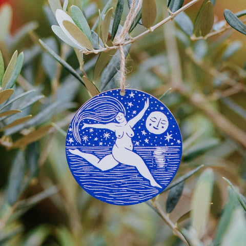 Blue decorative ornament with a woman and moon design hanging on a branch.
