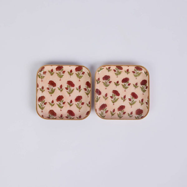 Two small square decorative coasters with rounded corners, featuring a repeating floral pattern in red and green on a light peach background, set against a plain light grey studio backdrop.