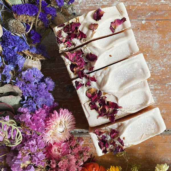 Bar of soap with dried flowers on a wooden surface surrounded by colourful flowers