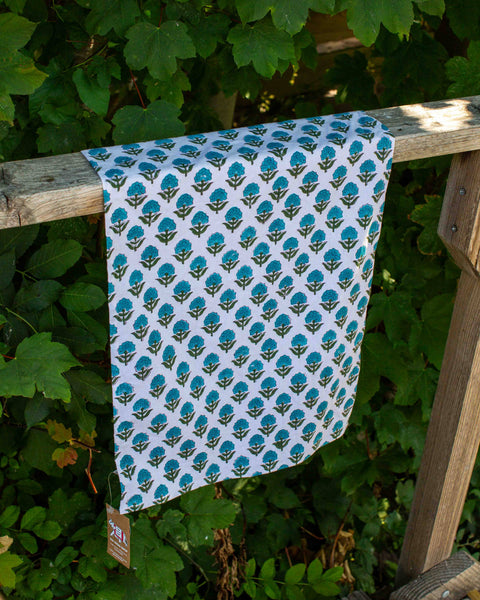 A white fabric tea towel with a repeating teal and olive green block print pattern draped over a rustic wooden railing, set against a background of lush green leaves.