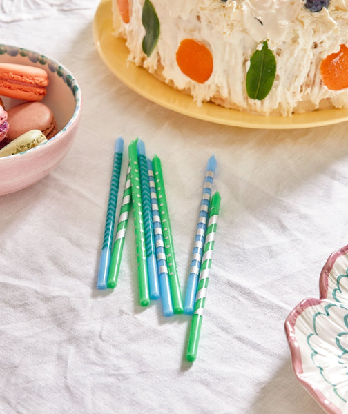 Colourful birthday candles on a white surface with a cake and macarons in the background.