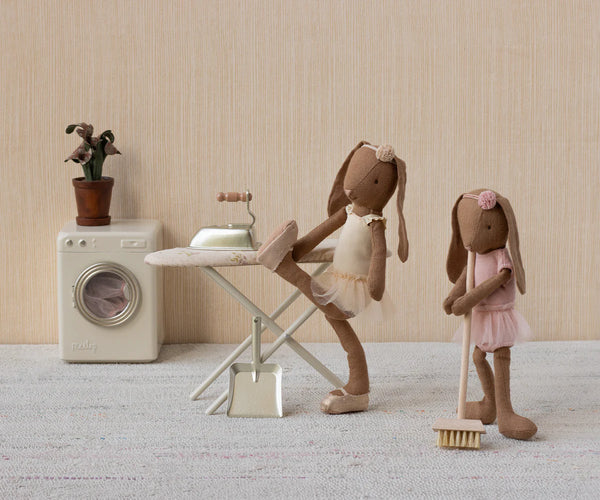 Two toy bunnies in a domestic setting with a washing machine, ironing board, and broom.