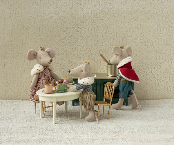 Three toy mice with a tea set on a small table against a patterned wall.