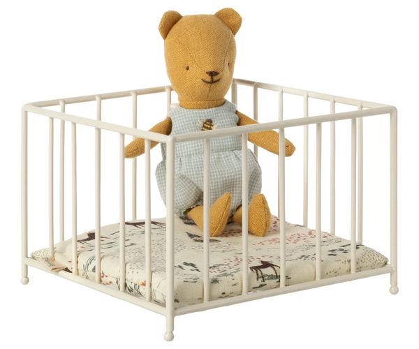 A small cream-coloured metal baby crib with a patterned mattress and a miniature teddy bear sitting inside, shown against a plain white background.