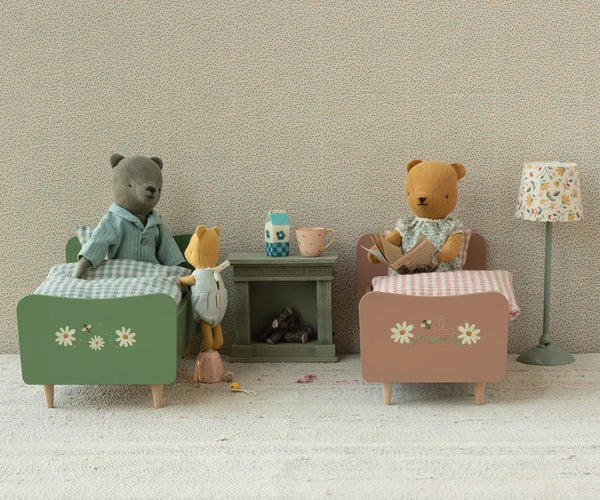 A miniature dollhouse-style bedroom scene with two teddy bears sitting in small beds, a small side table, a patterned lamp, and a textured wall background.