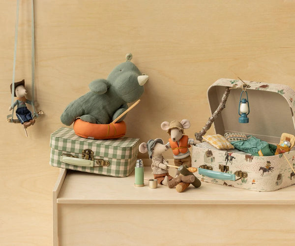 Toy mouse family with a suitcase and basket on a wooden shelf against a beige wall.