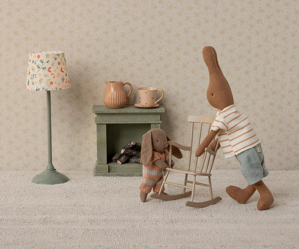 Two toy rabbits in a room with a lamp, fireplace and rocking chair
