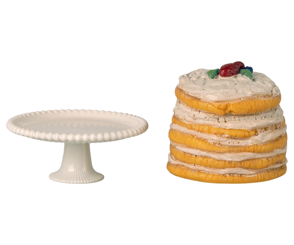 Stack of layered cookies with a cherry on top next to a white cake stand on a white background
