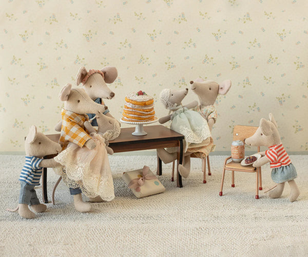 Stuffed animal mice gathered around a table with a cake in a room with floral wallpaper.