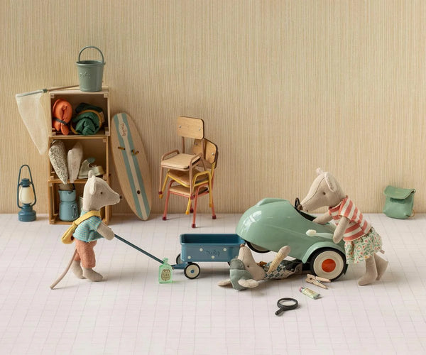 A styled miniature scene with small animal figurines and toy props arranged on a white floor against a beige wall. One figurine is pulling a blue wagon while another stands beside a mint green toy car, with additional beach-themed accessories and furniture in the background.