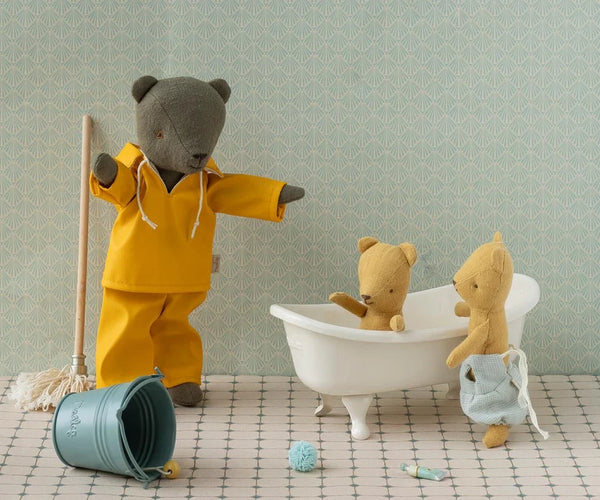 Three plush teddy bears arranged in a nursery-like scene: one standing bear in a yellow outfit holding a broom, one bear sitting in a white clawfoot tub, and one bear standing beside the tub holding a light blue cloth. A blue bucket lies on the floor, with a checkered floor and pale patterned wallpaper in the background.