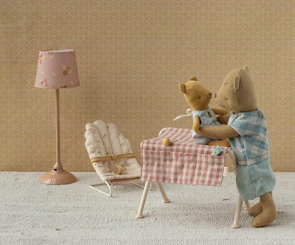 A styled miniature dollhouse interior featuring a teddy bear figure in a blue plaid shirt standing at a small table with a red-and-white checkered cloth, holding a smaller teddy bear in a light blue outfit. A pink floral floor lamp, a small chair, and patterned wallpaper fill the background.