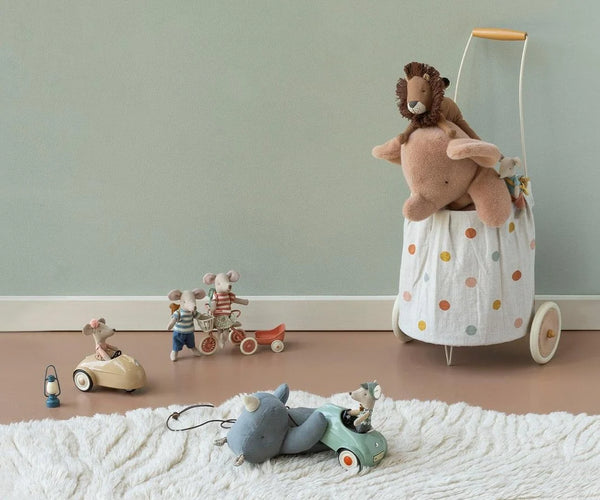 A styled children's play scene with a white fabric toy cart covered in pastel polka dots, holding a plush stuffed animal. Nearby are small wooden toy cars, a tricycle, and other soft play items on a white textured rug against a pale green wall.