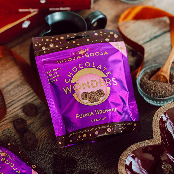 Purple package of Booja-Booja Chocolates Wonders Fudgie Brownie on a wooden surface with chocolate and other items.