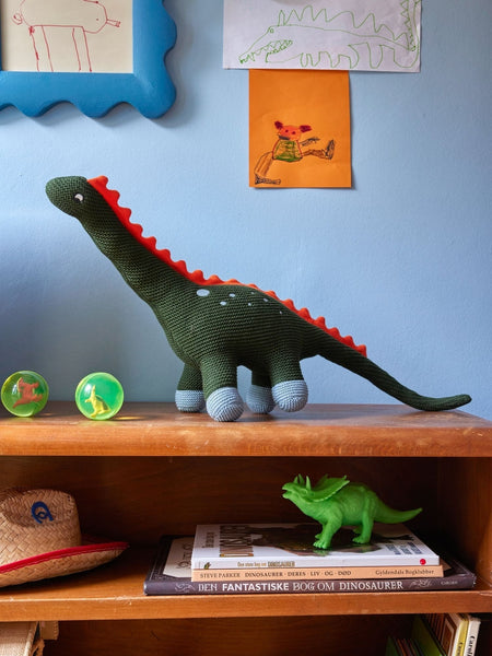Knitted dinosaur toy on a shelf with books and other items against a blue wall.