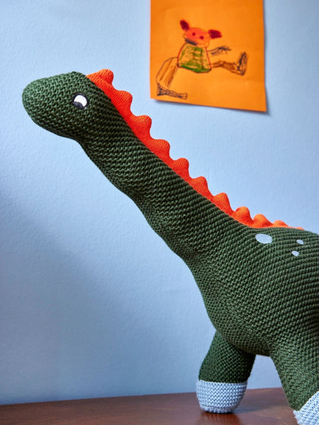Knitted dinosaur toy on a wooden surface with a light blue wall and a child's drawing in the background.