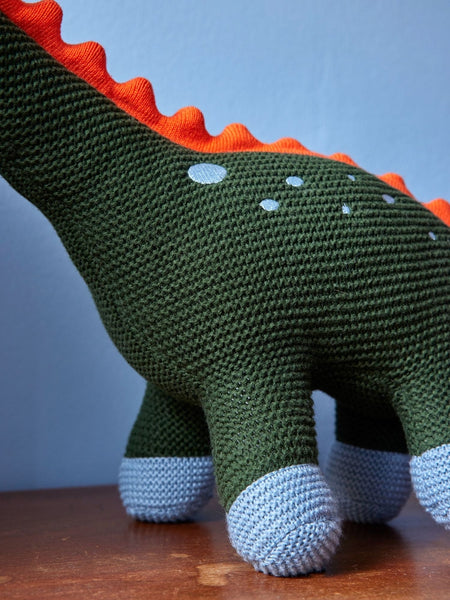 Knitted dinosaur toy with green body, orange spikes, and white feet on a wooden surface.